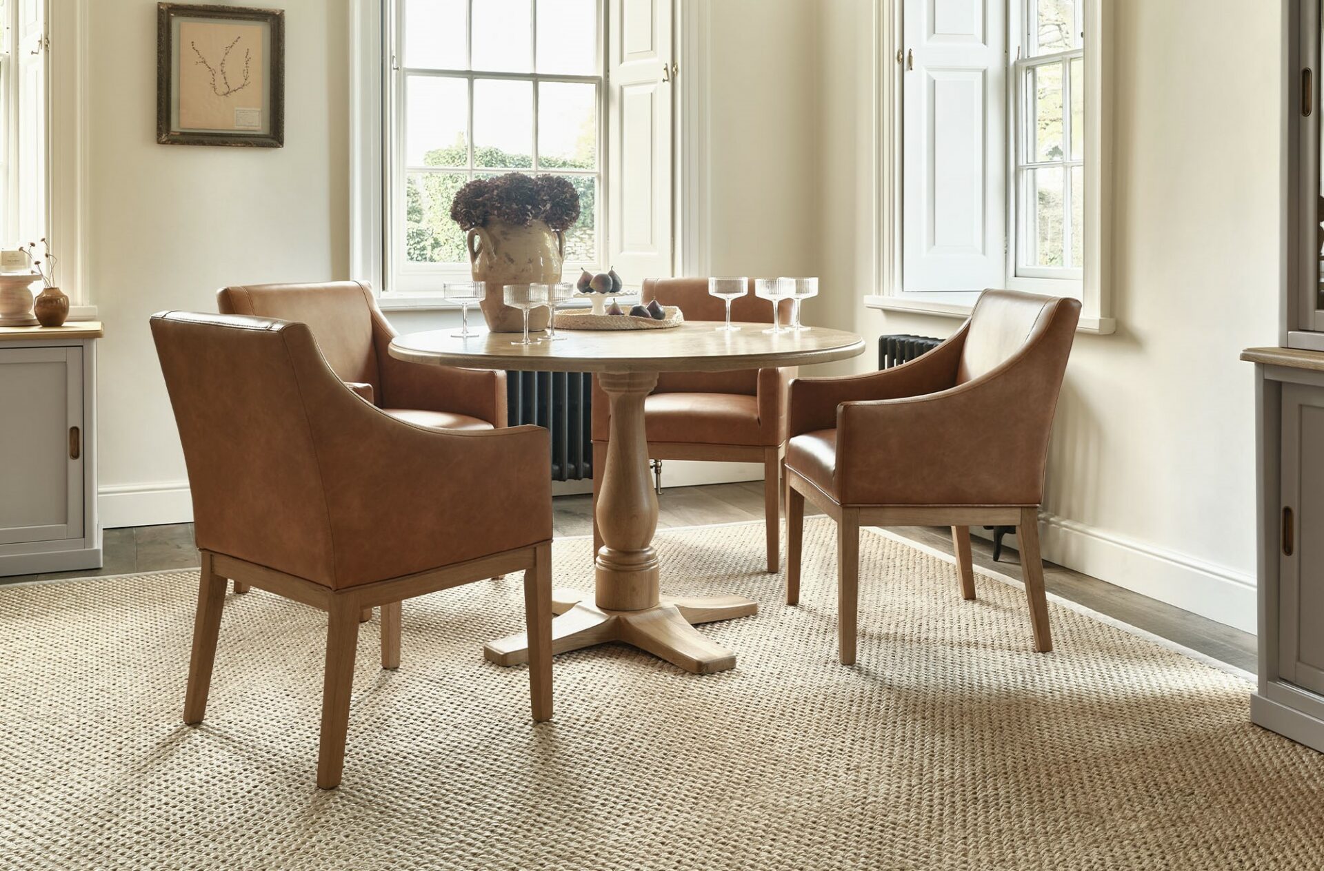 A dining table and dining chairs-dining room furniture-tan leather dining chair-round dining table-glassware-sisal rug-neutral walls-sideboard-dresser