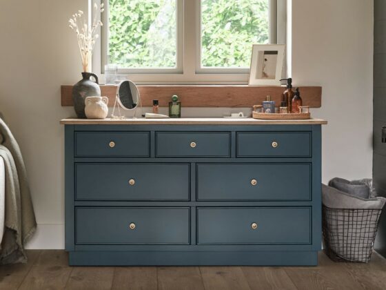 A chest of drawers and chair-bedroom furniture-wooden 7-drawer chest painted dark blue-neutral walls-upholstered accent chair-wooden shutters-wire storage basket