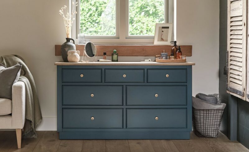 A chest of drawers and chair-bedroom furniture-wooden 7-drawer chest painted dark blue-neutral walls-upholstered accent chair-wooden shutters-wire storage basket