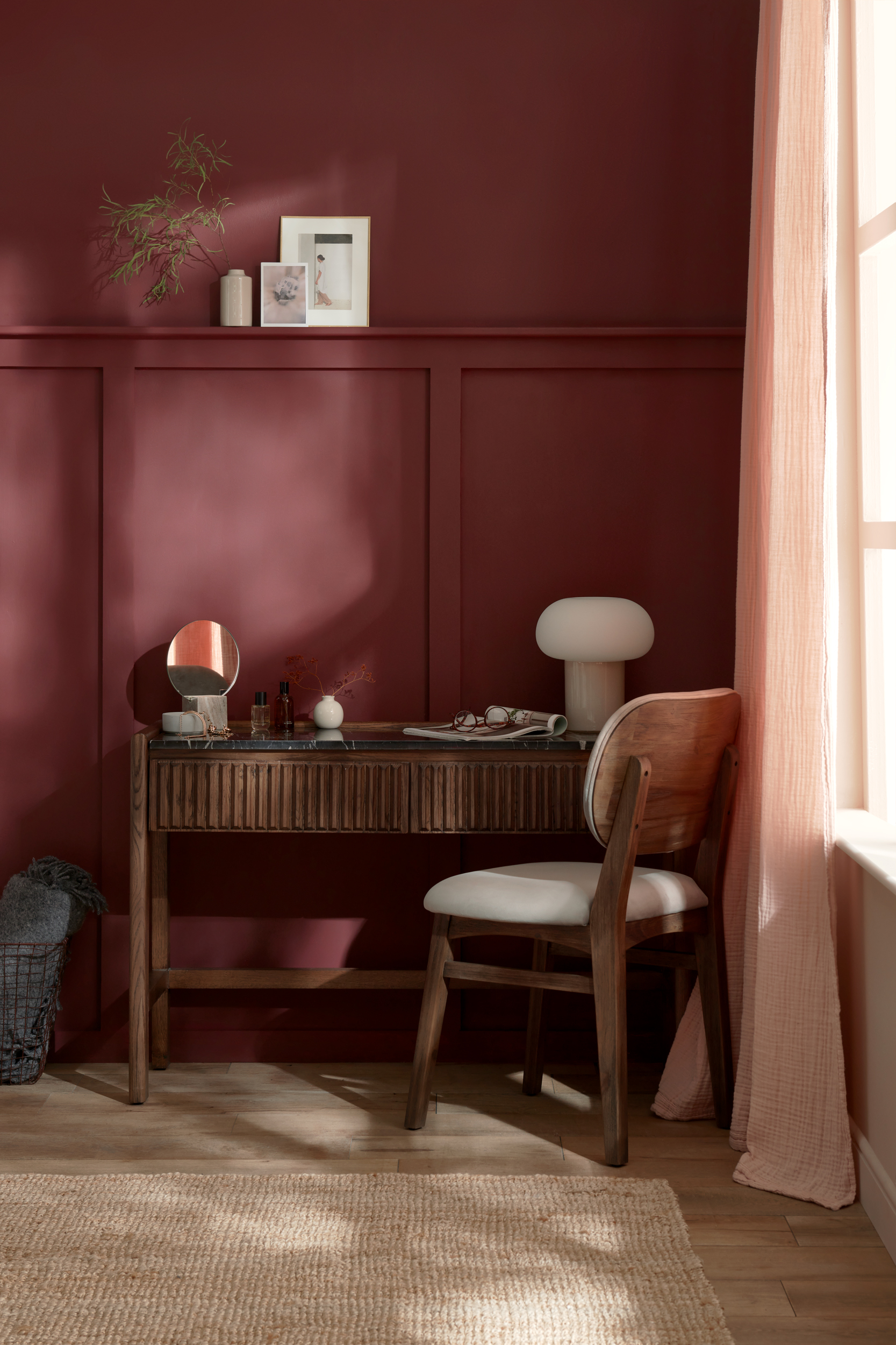 A dressing table and chair-bedroom furniture-burgundy walls-cream lamp-dark wooden dressing table-upholstered chair-wall panelling-pink curtain-jute rug