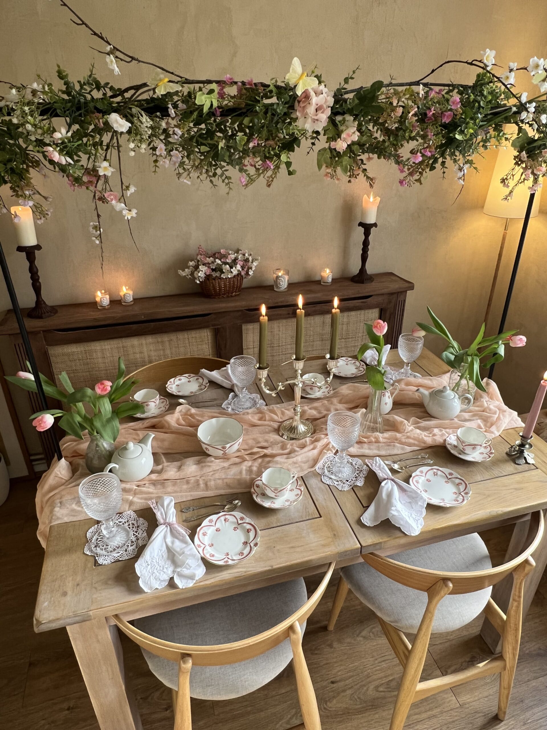 A dining table and chairs-dining room-vintage-style china-delicate glassware-white linen napkins-soft pink table runner-tulips in vases-lit candles-a floral arrangement with greenery and pastel flowers-wooden sideboard